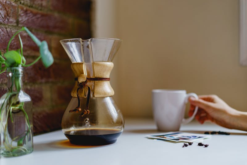 Café dans une Chemex sur une table blanche à côté d'un mur de briques et une main tenant une tasse à café blanche.