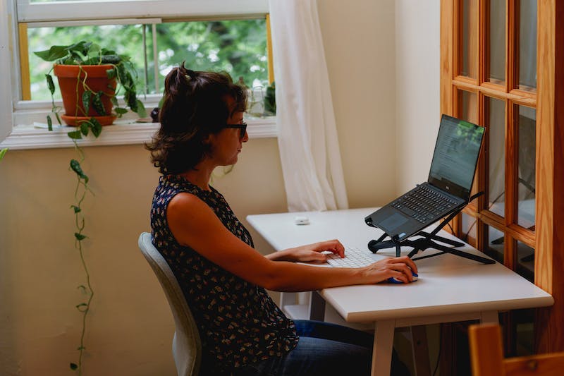 Femme utilisant un support pour ordinateur portable avec un ordinateur portable sur un petit bureau blanc compact dans un bureau à domicile dans un appartement.