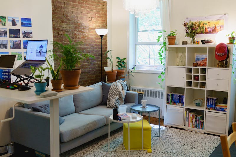 Installation d'un bureau debout dans un petit appartement avec un canapé, un ottoman, une bibliothèque et un mur en briques apparentes.