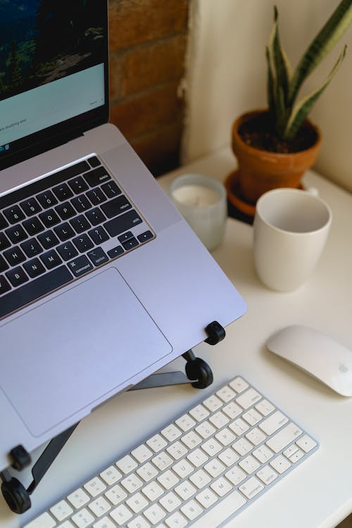 MacBook Pro sur un support d'ordinateur portable dans une configuration de bureau à domicile sur un bureau blanc avec un clavier externe, une souris Apple, une bougie, une plante et une tasse.