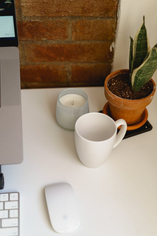 Bureau de travail blanc avec tasse blanche, petite bougie, plante en pot et souris sans fil blanche contre un mur de briques apparentes.