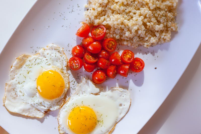 Assiette rectangulaire blanche avec deux œufs au plat, des tomates raisin sautées et du couscous parsemé d'herbes.