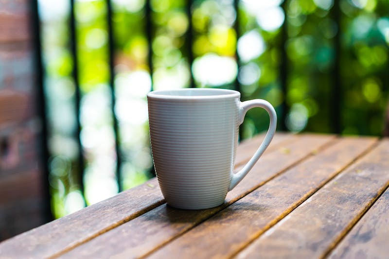 Tasse à café blanche posée sur une table d'extérieur en bois avec des arbres verts en arrière-plan.