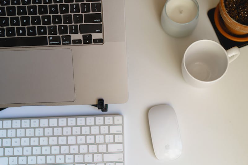 Vue à plat d'un bureau à domicile avec un ordinateur portable MacBook Pro, un clavier sans fil, une souris sans fil, une bougie, une tasse et une plante sur une surface de bureau blanche et fine.
