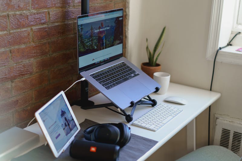 Mur de briques apparentes dans un salon avec un bureau à domicile, un ordinateur portable argenté sur un support noir, un clavier sans fil blanc, une souris sans fil et des écouteurs bluetooth noirs.