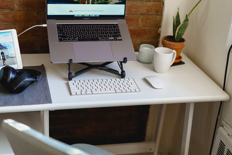 Petit bureau blanc mince pour le bureau à domicile, équipé d'un ordinateur portable, d'un clavier sans fil, d'une chaise de bureau vert clair et d'une souris sans fil blanche, contre un mur de briques.