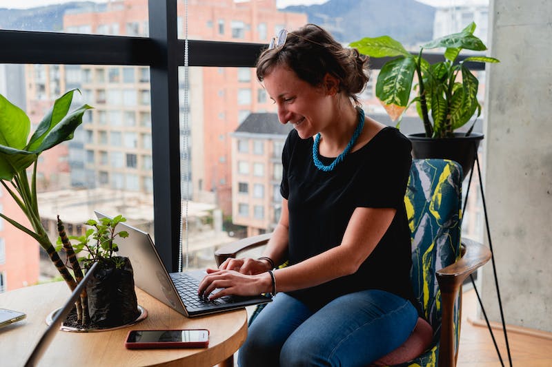 Femme travaillant à distance sur un ordinateur portable à une table ronde en bois dans un espace de coworking avec des plantes.