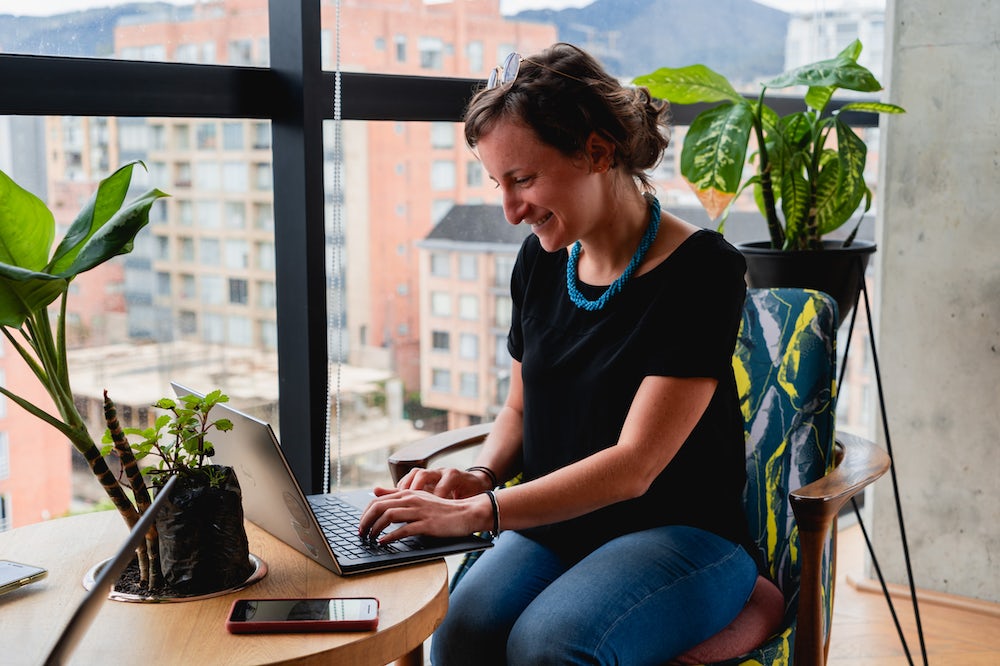 Femme travaillant à distance à partir d'un ordinateur portable à une table ronde en bois dans un espace de coworking avec des plantes.