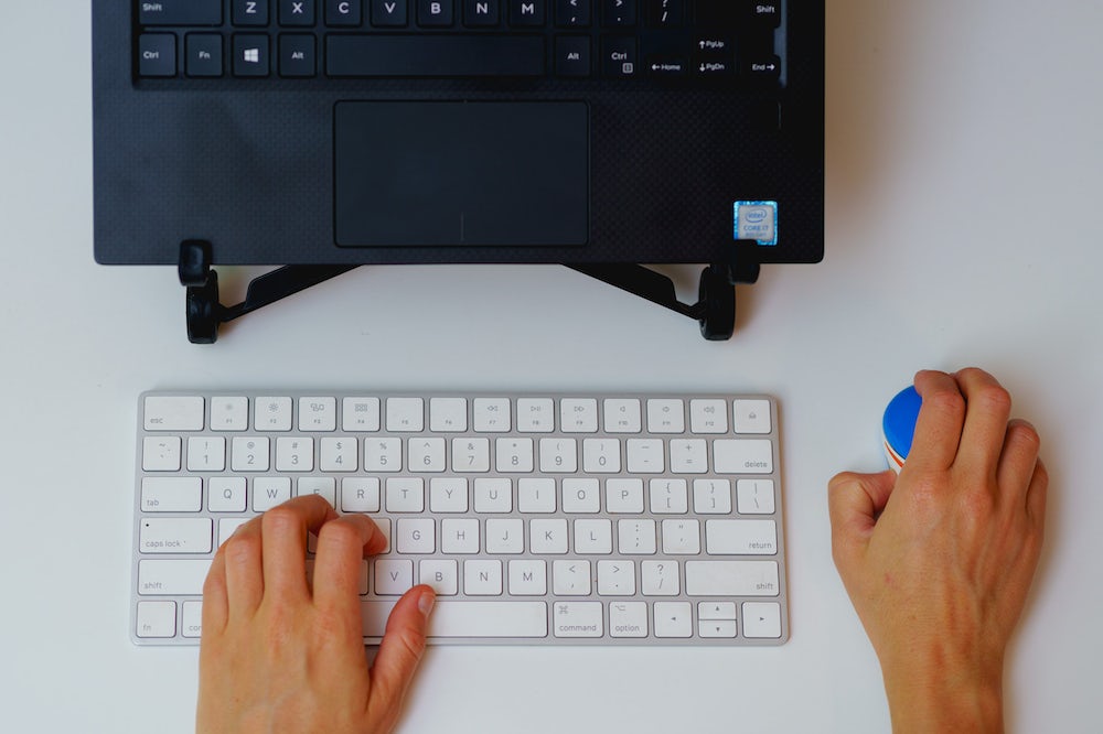 Femme utilisant une petite souris sans fil avec un clavier apple magic et un ordinateur portable sur un support dans une configuration de bureau à domicile.