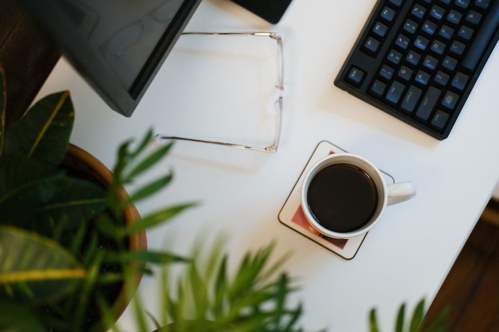 Mise en page plate d'un bureau à domicile avec des lunettes transparentes, une tasse blanche remplie de café, des plantes vertes et un clavier noir.
