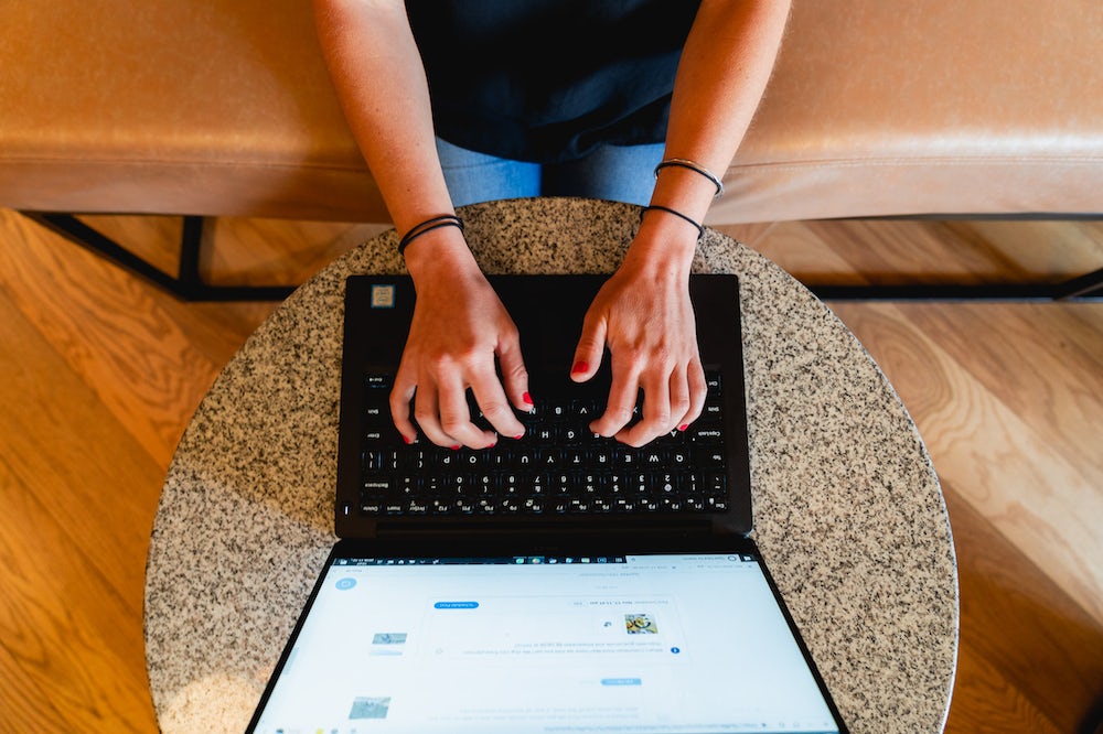 Femme assise sur un banc beige travaillant à distance sur un ordinateur portable à une petite table circulaire en granit.