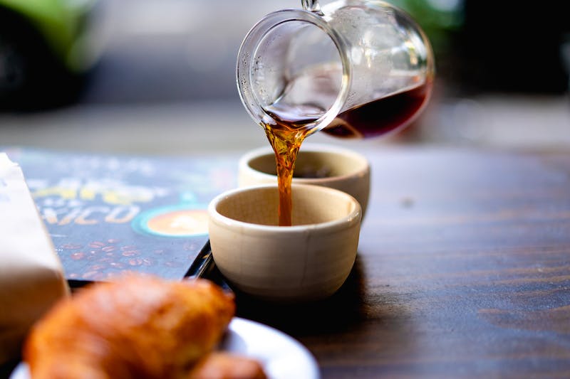 Versement d'un filet de café noir d'une carafe en verre sur une table en bois dans une petite tasse blanche.