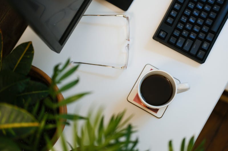 Flat lay d'un bureau à domicile avec des lunettes transparentes, une tasse blanche remplie de café, des plantes vertes et un clavier noir.